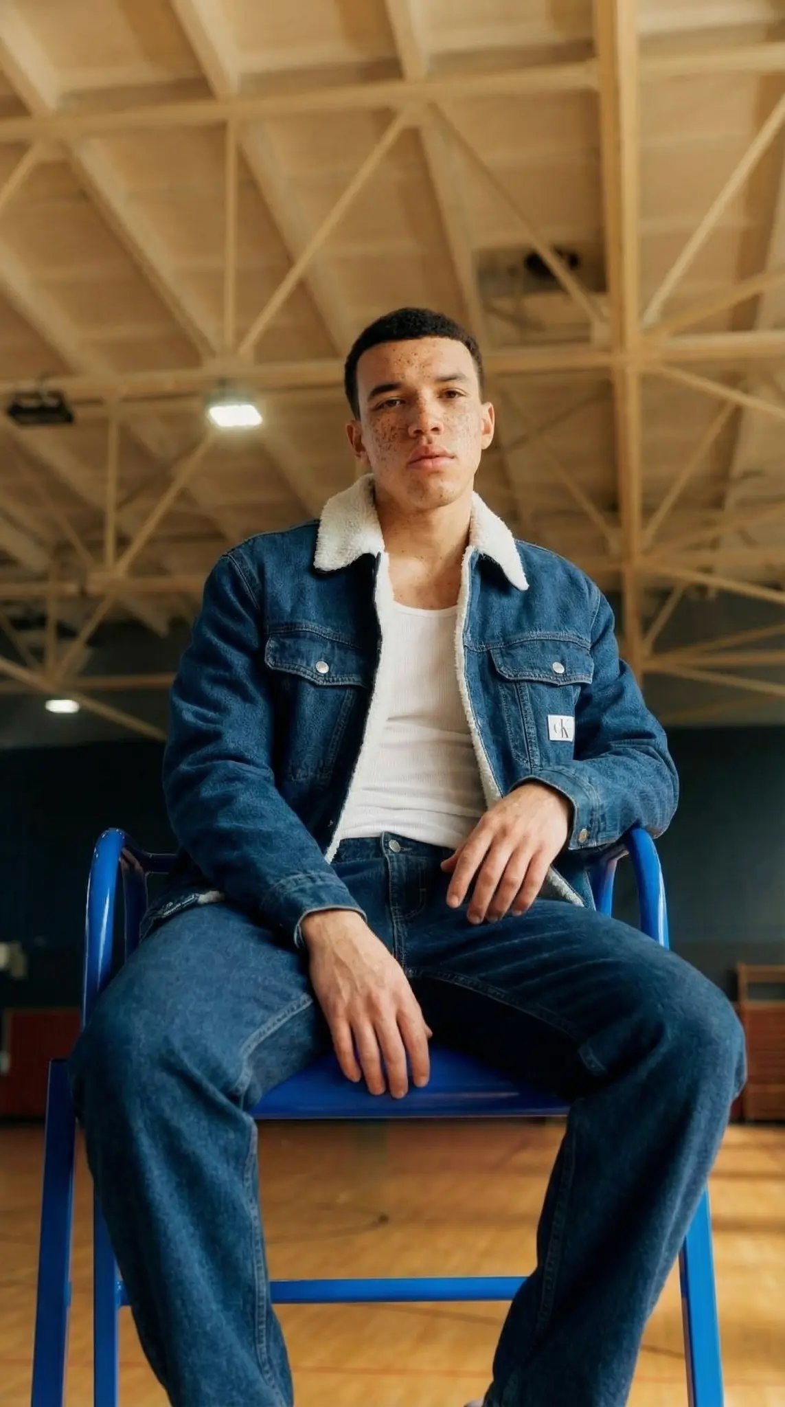 Calvin Klein denim campaign featuring model in sherpa-lined denim jacket sitting on blue chair in gymnasium setting